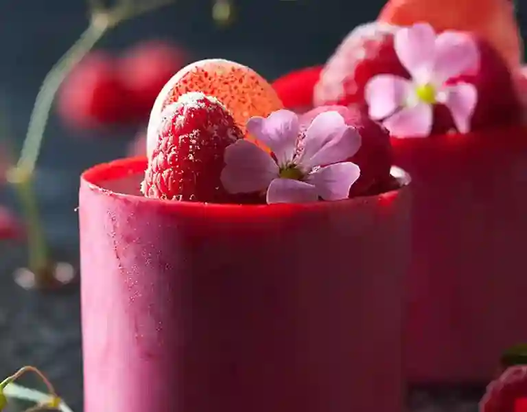 A refreshing raspberry beverage served in a tall glass with ice cubes, showcasing vibrant red hues and garnished with fresh raspberries on the rim